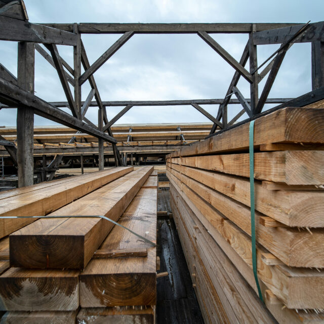 Stacks of wooden planks in a lumber yard under a cloudy sky with metal framework overhead.