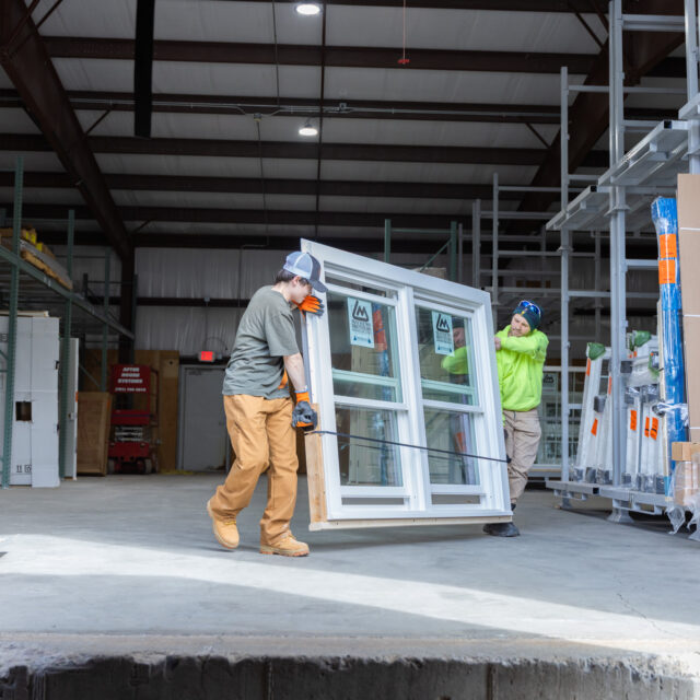 Workers in safety gear carrying a large window frame inside a warehouse with construction materials in the background.