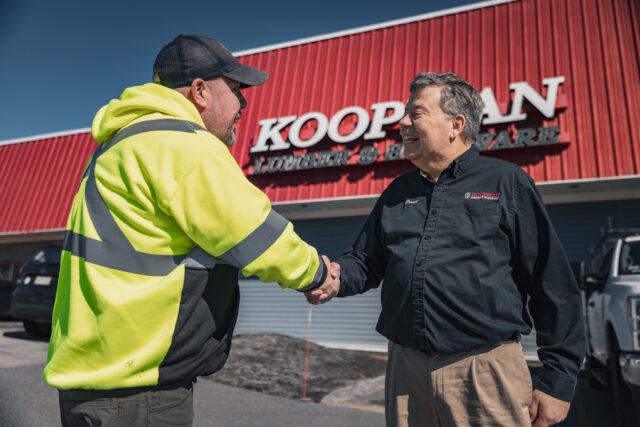 Two men shaking hands in front of a Koopman Lumber and Hardware store, both wearing jackets outside.