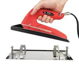 Hand holding a red electric tool above a metal base with clips, possibly for craft or repair work.