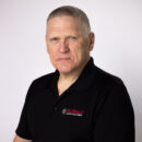 Man in a black polo shirt with a company logo on a plain white background.