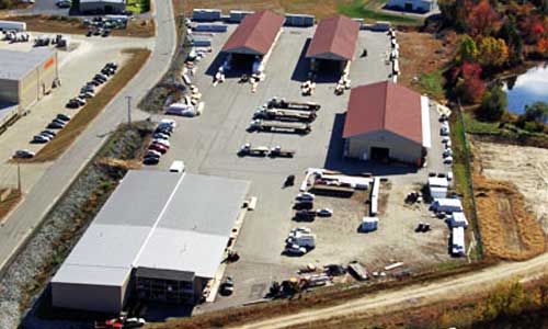 Aerial view of industrial warehouses with parked vehicles and storage areas in a rural landscape.