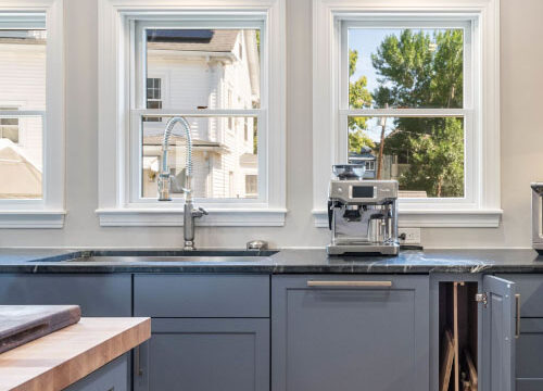 Modern kitchen with grey cabinets, natural light from large windows, and a coffee machine on the counter.