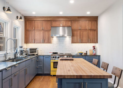 Modern kitchen with wooden cabinets, blue island, stainless steel appliances, and wooden stools under bright ceiling lights.