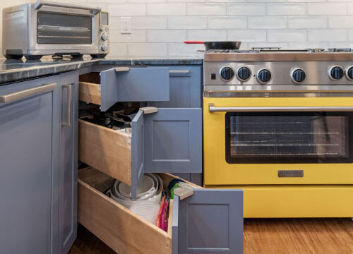 Modern kitchen with open blue cabinets, yellow oven, and wooden floor. Small toaster oven on the counter.