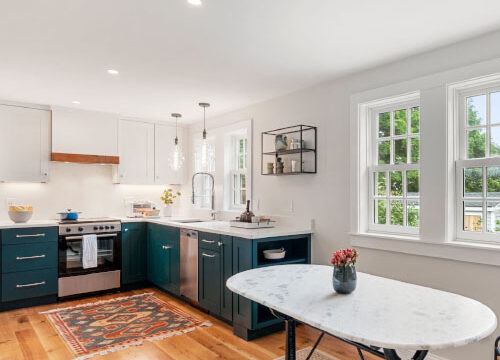 Bright kitchen with teal cabinets, marble countertops, and a white table on hardwood flooring, featuring large windows.