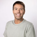 Man smiling in a light grey textured shirt against a plain background.