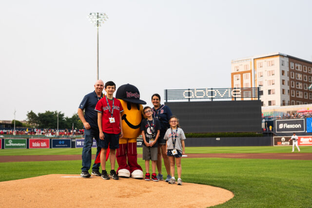 Group posing with a mascot on a baseball field, stadium and lights in the background.