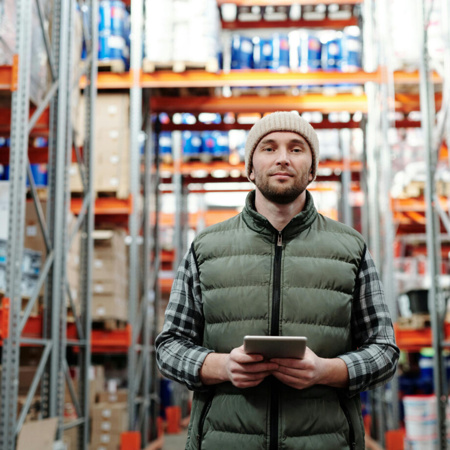 Warehouse worker in a vest holds a tablet, standing among tall shelves stocked with various boxes and packages.