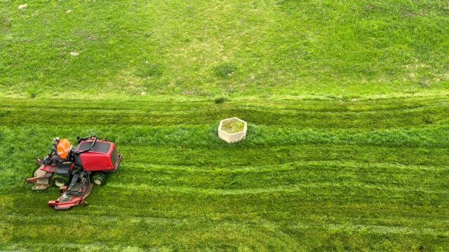 A person in an orange vest rides a mower in a lush green field.