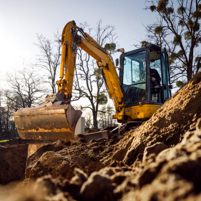 Yellow excavator digging earth at a construction site on a sunny day, surrounded by trees and piles of dirt.