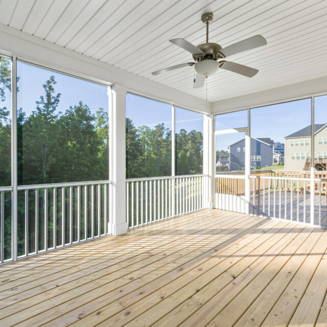 Screened-in porch with wooden flooring, ceiling fan, and view of trees and houses in the background.