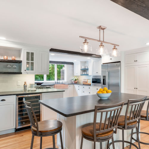 The picture depicts a kitchen with an island and four high chairs. There is a bowl of oranges on the table and a stainless steel refrigerator in the background.