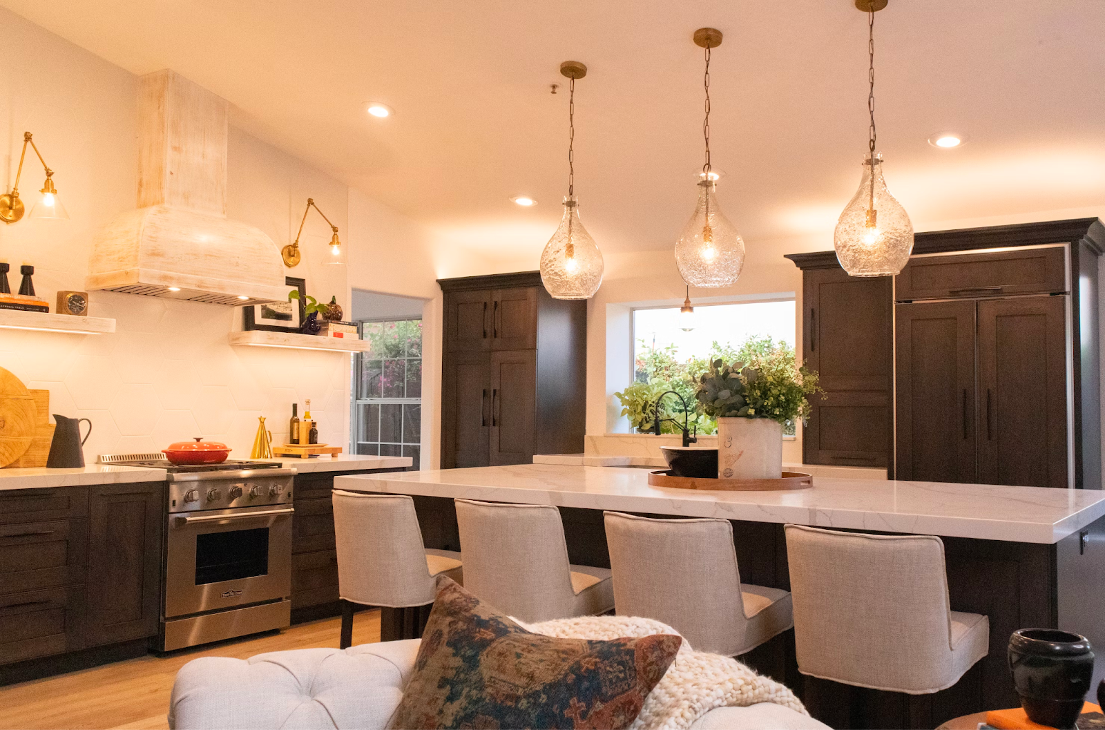 The picture depicts a kitchen with three light fixtures and four upholstered high chairs. The cabinets are dark wood and the lighting in the house is warm.