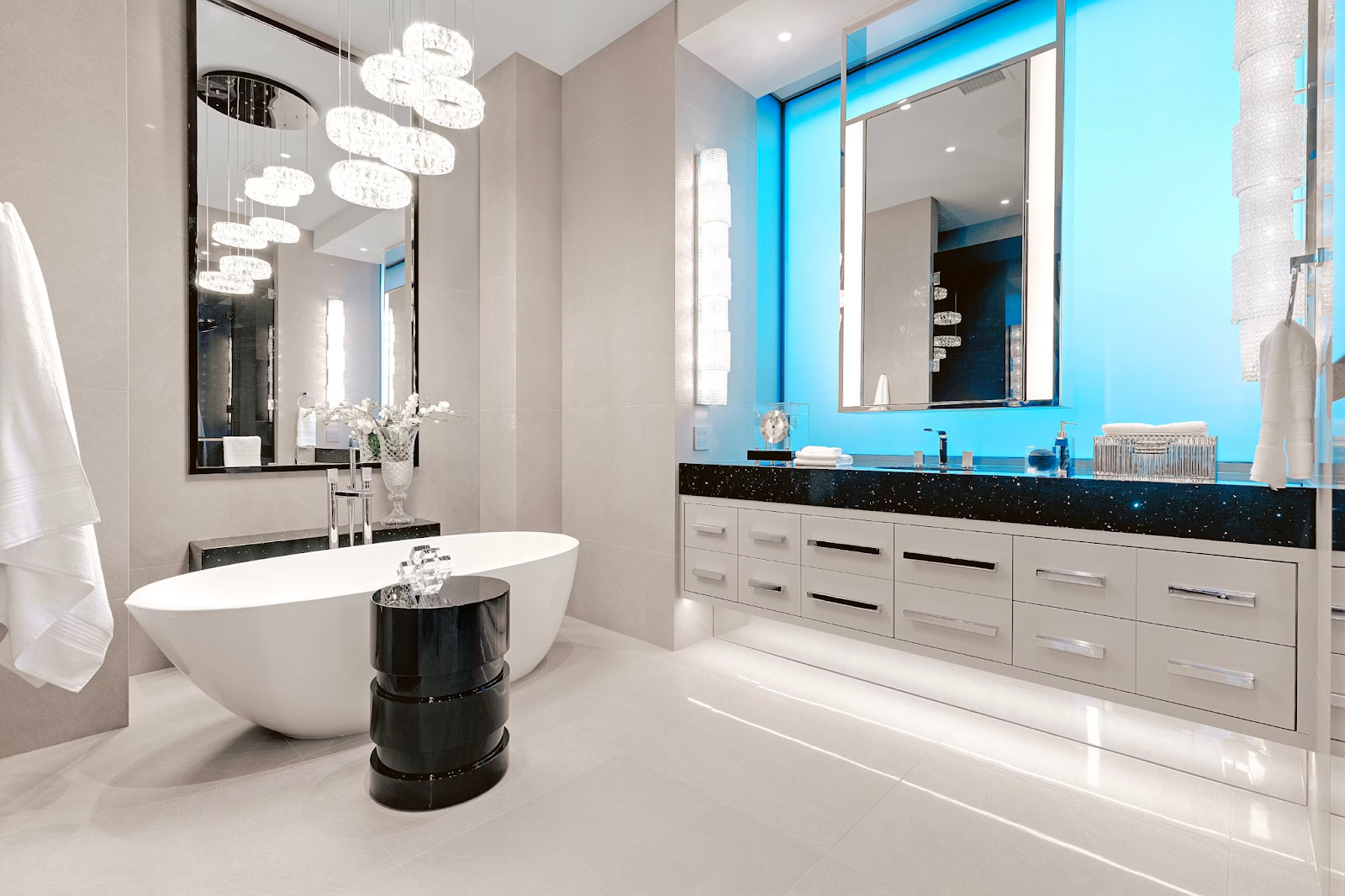 The picture depicts a modern bathroom with a stand-alone bathtub and a shiny, black side table. The bathroom sink has marble countertops and a large mirror with blue LED lights.
