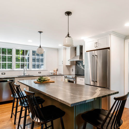 The picture depicts a kitchen with four high chairs around a kitchen island. There is a stainless steel refrigerator and large sink as well.