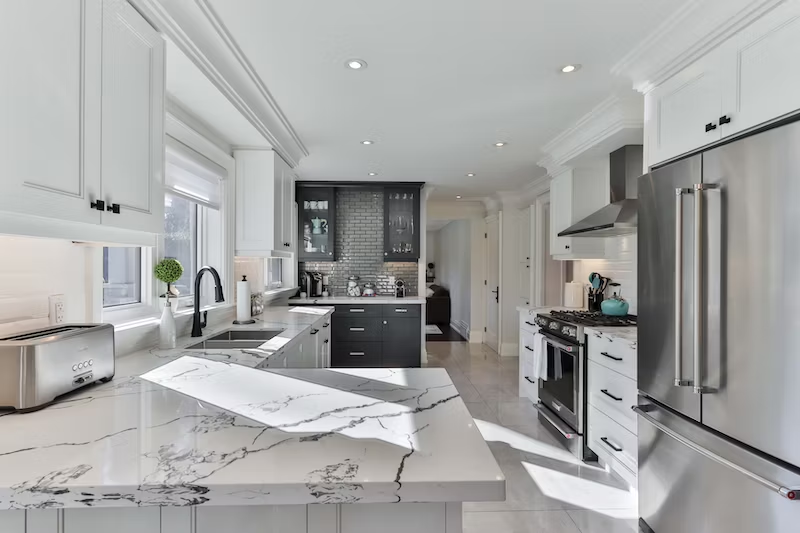 The picture depicts a white kitchen with marble countertops. There is a stainless steel fridge to the right and black cabinets in the back.