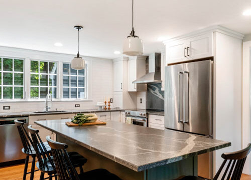 Modern kitchen with stainless steel appliances, an island with a wooden countertop, and black chairs under pendant lighting.