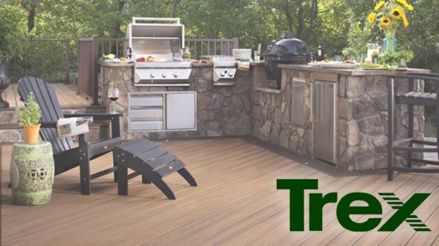 Outdoor kitchen on a wooden deck with grill, cabinets, and seating surrounded by greenery.