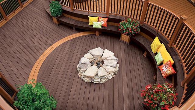 wooden deck with circular stone fire pit, surrounded by curved benches with colorful pillows and potted red flowers