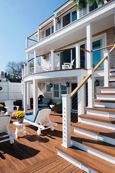Two-story white house with wooden deck, outdoor seating, white chairs, and potted flowers on a sunny day.