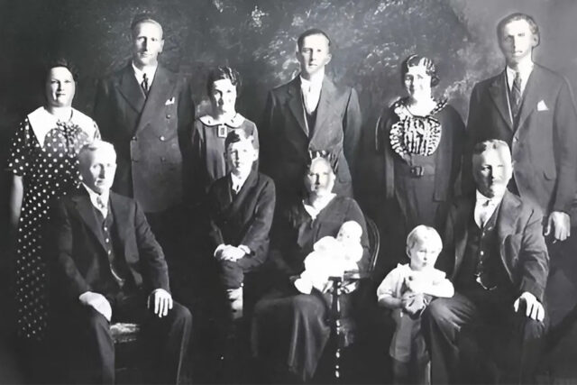 Black and white vintage family portrait with three generations posing in formal attire against a dark backdrop.