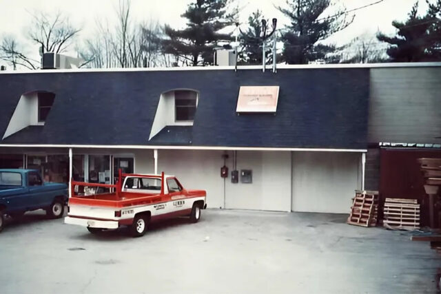 Red pickup truck parked outside a two-story building with trees in the background, overcast sky.