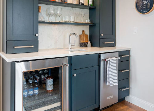 Home bar with dark cabinets, countertop sink, and stocked mini fridge. Wall-mounted clock on the right.