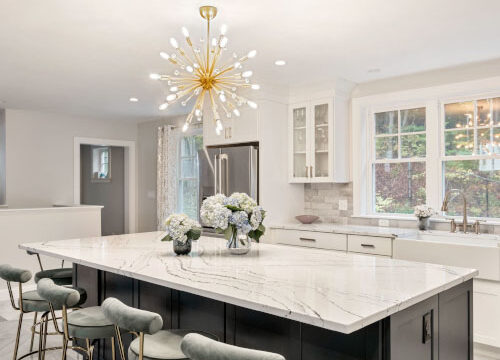 Modern kitchen with marble island, gold chandelier, bar stools, and floral arrangement.