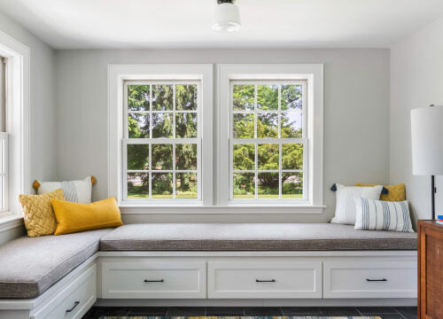 Cozy window nook with cushions and throw pillows, surrounded by white drawers and accented by a colorful rug.