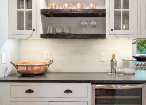Modern kitchen with white cabinets, glass shelves, and a small wine fridge. Copper pot and glasses on counter.