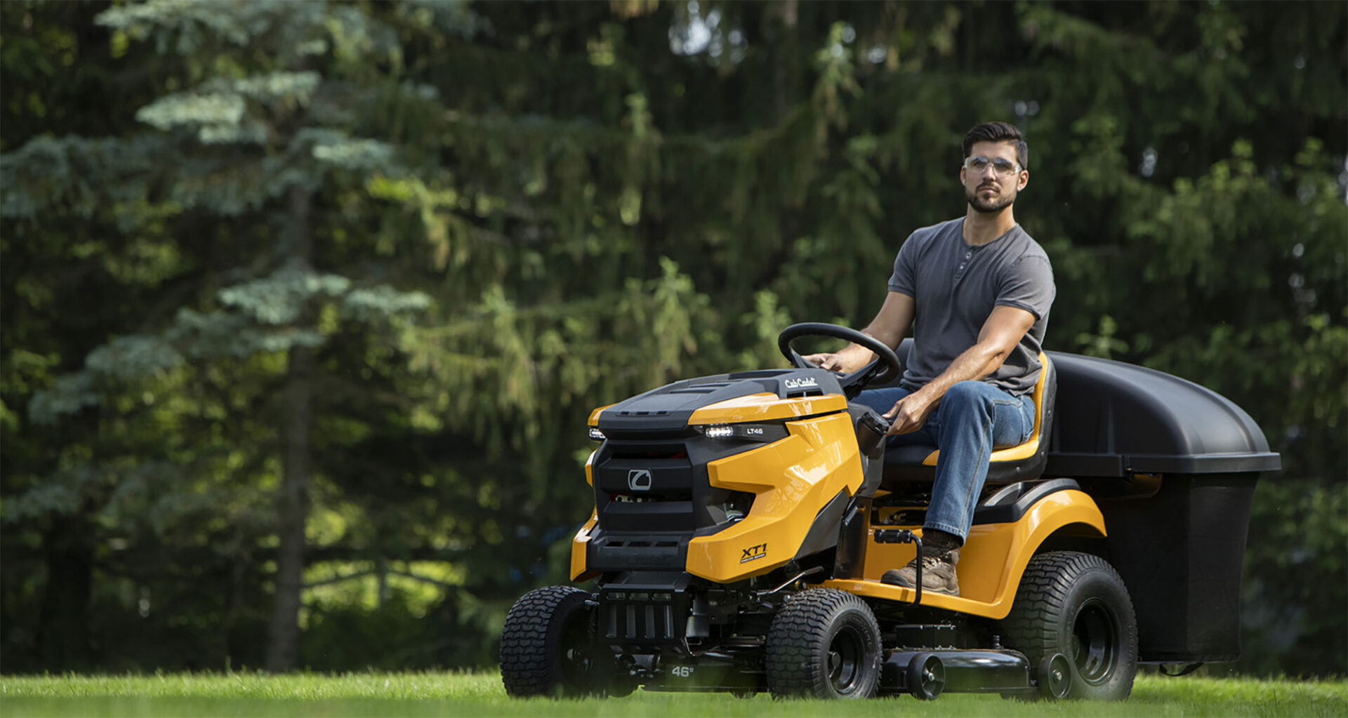Man mowing a lawn with a yellow riding lawnmower, surrounded by green trees in a sunny garden.
