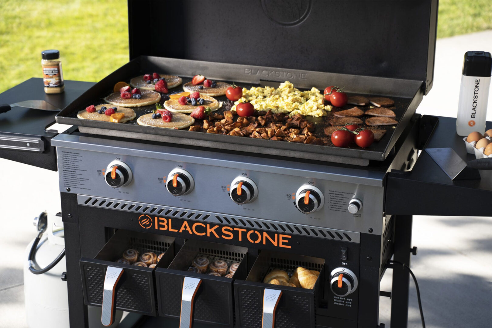 Outdoor griddle with pancakes, berries, scrambled eggs, tomatoes, and sausage, ready for breakfast cooking.