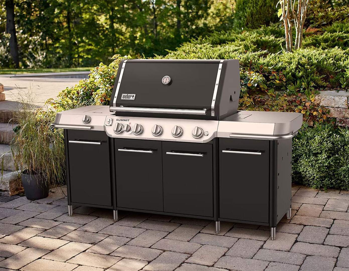 Outdoor gas grill with six burners, black finish, and stainless steel accents, placed on a stone patio with greenery behind.