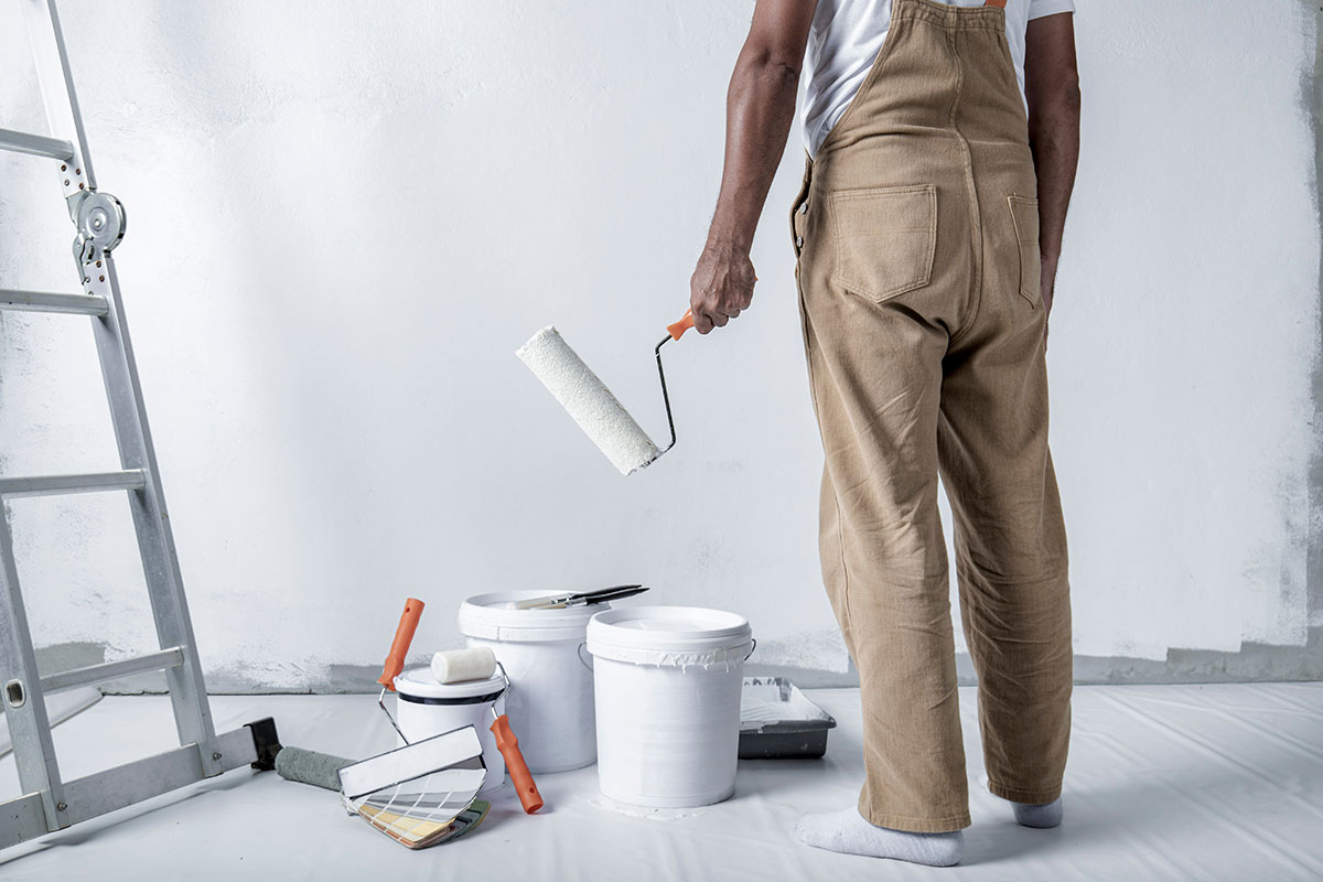 Person in overalls holding a paint roller, standing next to paint buckets and brushes on a floor covered with a white sheet.